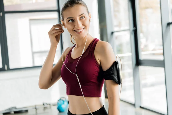 Beautiful smiling sportswoman in smartphone armband putting on earphones at gym — Stock Photo