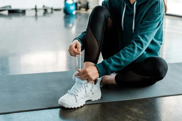 Vista recortada de la deportista atando cordones en zapatos de entrenamiento mientras está sentado en la alfombra de fitness en el gimnasio - foto de stock