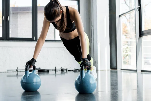 Strong sportswoman in weightlifting gloves doing plank exercise on kettlebells at gym — Stock Photo