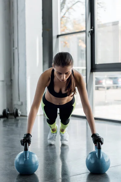 Sportiva concentrata in guanti di sollevamento pesi facendo esercizio di tavola su kettlebells presso il centro sportivo — Foto stock