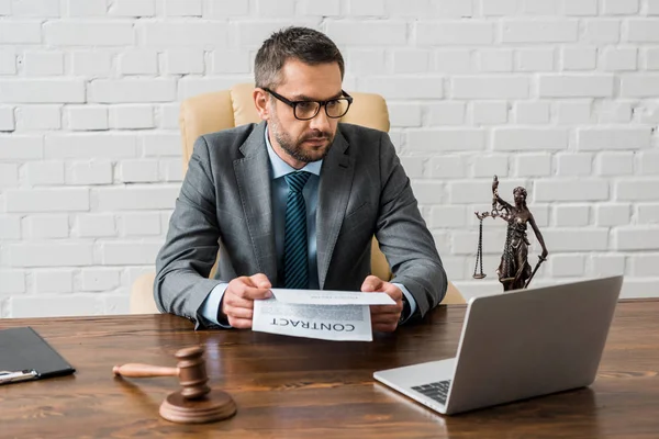 Serious male lawyer holding contract and looking at laptop in office — Stock Photo