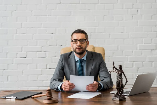 Juez serio en las gafas que trabajan con papeles y mirando a la cámara en la oficina - foto de stock