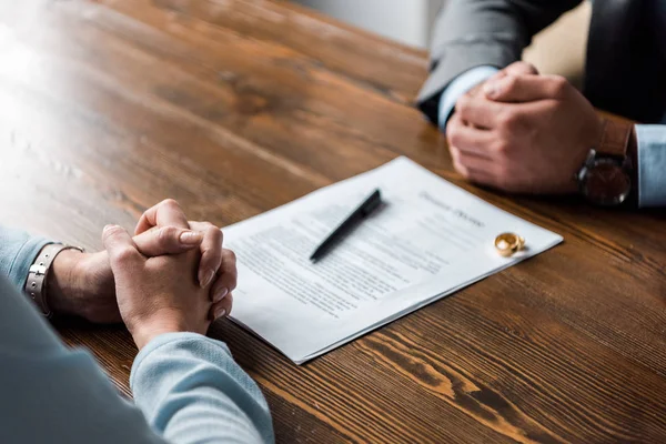 Vista parcial de las manos de abogado y cliente, decreto de divorcio y anillos de boda en la mesa - foto de stock