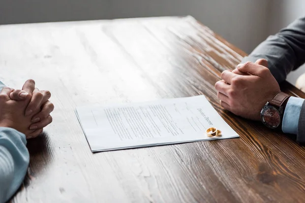 Tiro recortado de mãos de advogado e cliente, decreto de divórcio e alianças de casamento na mesa — Fotografia de Stock