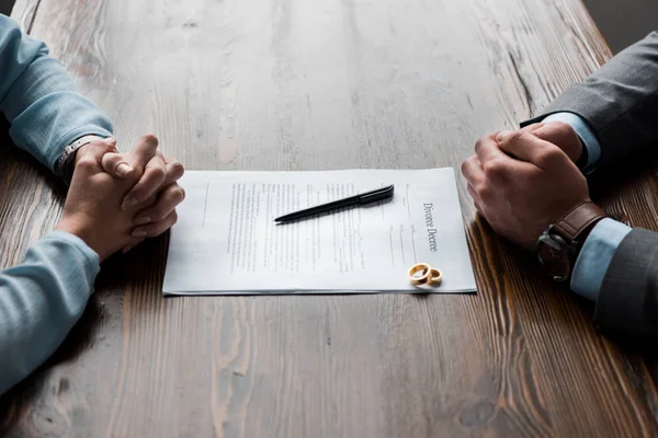 Recortado tiro de abogado y cliente sentado en la mesa con el decreto de divorcio y anillos de boda - foto de stock