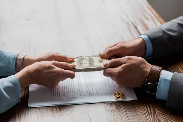 Vista parcial de abogado y cliente con billetes en dólares por encima del decreto de divorcio con anillos de boda - foto de stock