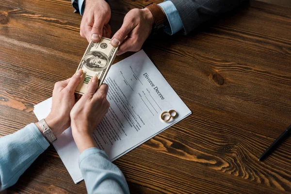 Vista superior parcial de la mujer dando dinero a abogado sobre el decreto de divorcio con anillos de boda - foto de stock