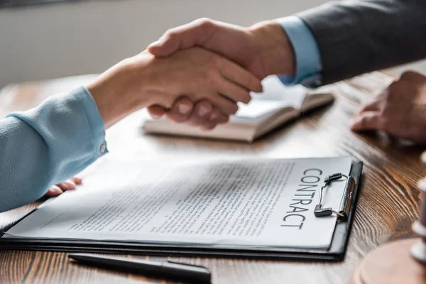 Close-up view of clipboard with contract and lawyer with client shaking hands behind — Stock Photo