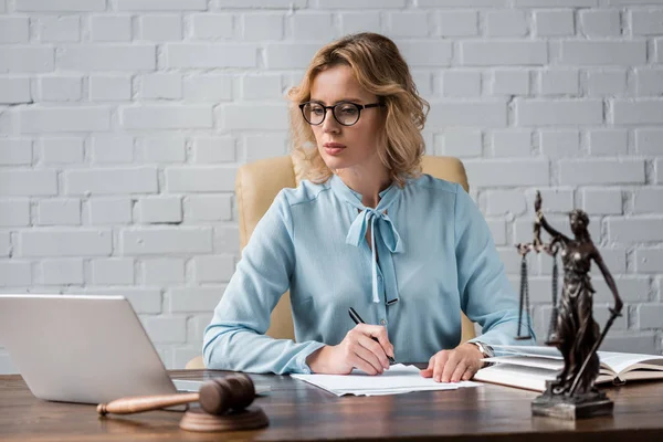 Juiz feminino grave em óculos trabalhando com papéis e usando laptop no local de trabalho — Fotografia de Stock