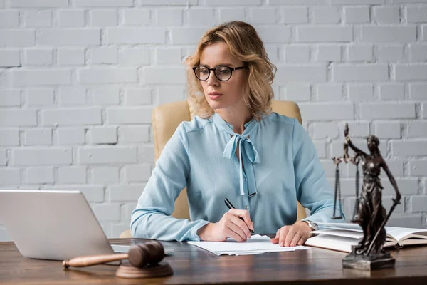 Juez femenino serio en las gafas que trabajan con papeles y el uso de la computadora portátil en el lugar de trabajo - foto de stock