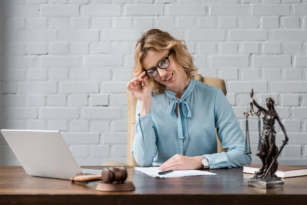 Beautiful female lawyer in eyeglasses smiling at camera while sitting at workplace — Stock Photo