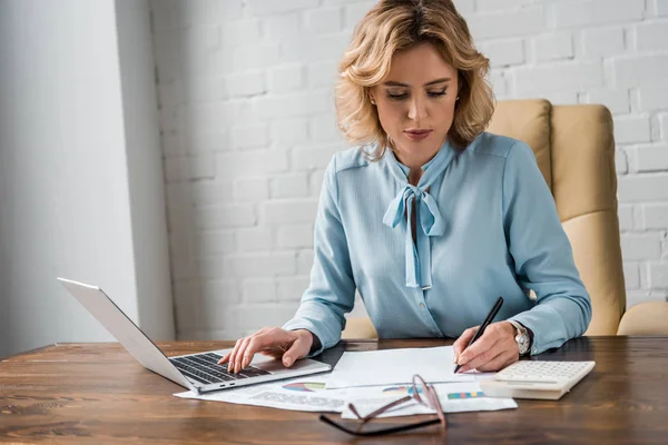 Fokussierte Geschäftsfrau, die im Büro mit Papieren und Laptop arbeitet — Stockfoto