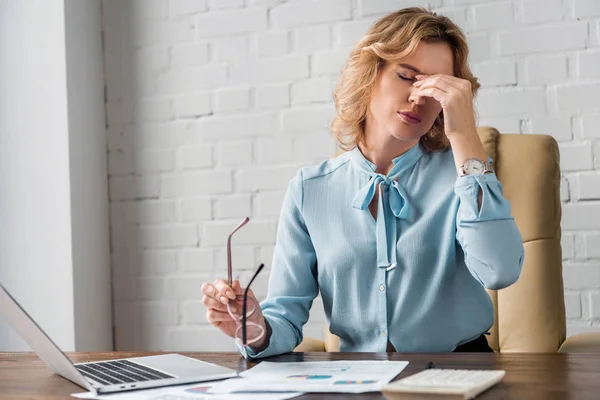 Mujer de negocios cansada sosteniendo anteojos y frotando puente nasal en el lugar de trabajo - foto de stock