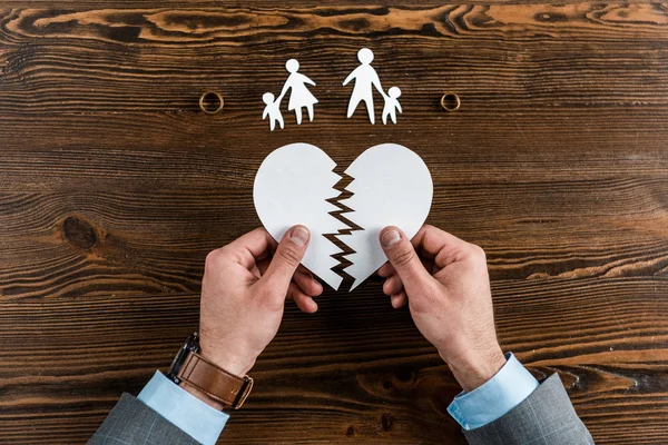 Plan recadré de l'homme tenant cœur déchiré avec la famille et les bagues de fiançailles sur une table en bois, concept de divorce — Photo de stock