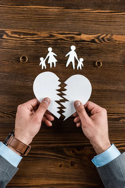 Tiro cortado do homem segurando o coração lacrimogêneo com a família e anéis de noivado na mesa de madeira, conceito de divórcio — Fotografia de Stock