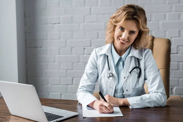 Souriant femme médecin écriture document sur le lieu de travail dans le bureau et regarder la caméra — Photo de stock