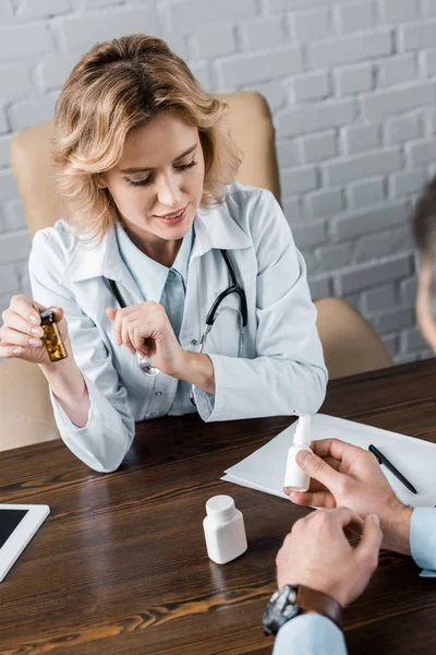 Vista ad alto angolo di attraente medico femminile che mostra vaso di pillole al paziente in ufficio — Foto stock