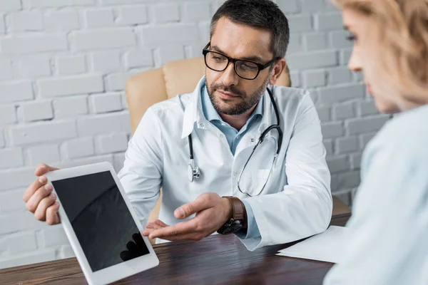 Médico bonito mostrando tablet com tela em branco para o paciente no escritório — Fotografia de Stock