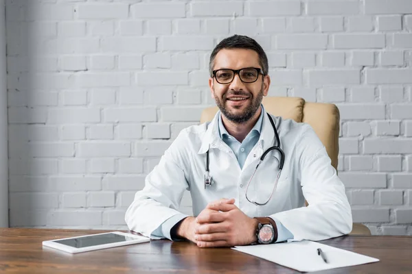 Handsome happy doctor looking at camera while sitting at workplace at office — Stock Photo