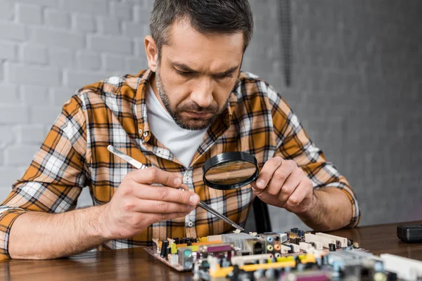 Portrait en gros plan de l'ingénieur en électronique concentré avec pince à épiler et loupe réparer carte mère — Photo de stock