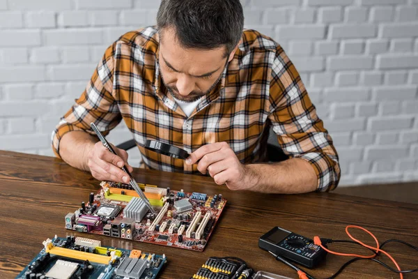 Ingénieur en électronique concentré avec pince à épiler et loupe réparer carte mère — Photo de stock