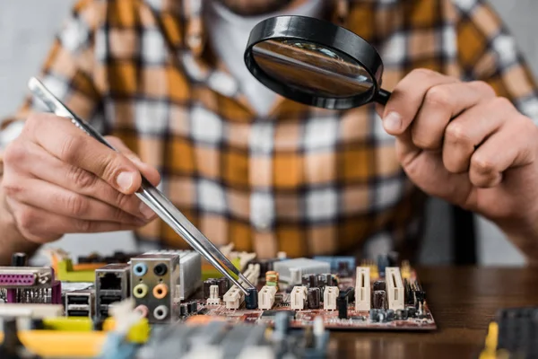 Cropped shot of computer engineer with tweezers and magnifying glass repairing motherboard — Stock Photo