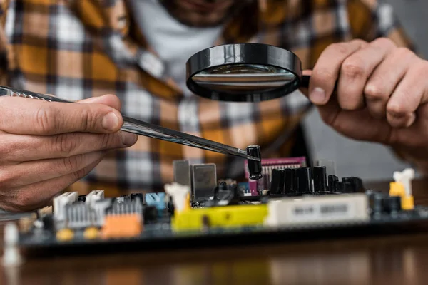 Cropped shot of electronics engineer with tweezers and magnifying glass repairing motherboard — Stock Photo