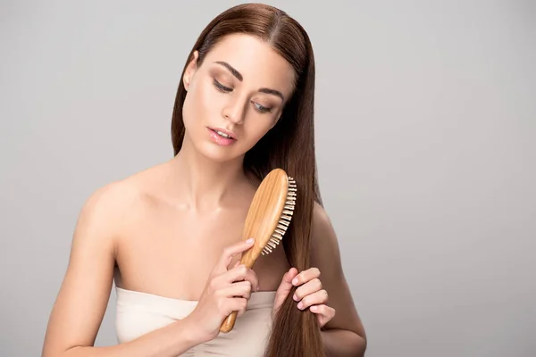 Atractiva chica morena peinando el cabello con cepillo de pelo de madera natural, aislado en gris - foto de stock
