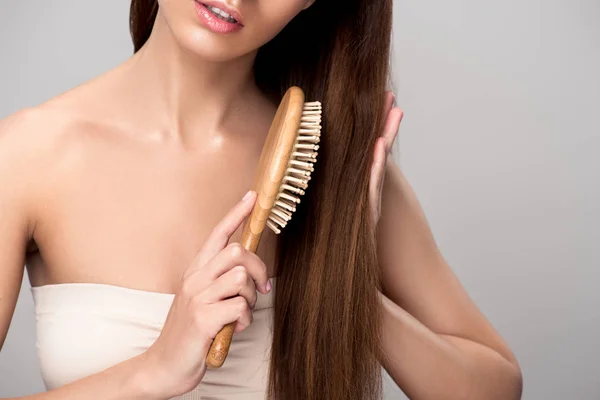 Vue recadrée de la femme peigner les cheveux avec une brosse à cheveux en bois, isolé sur gris — Photo de stock