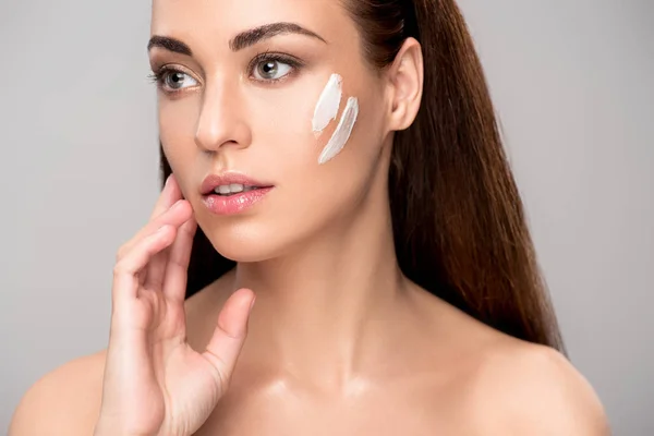 Attractive young woman applying cream on face, isolated on grey — Stock Photo