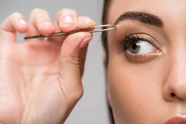 Close up of young woman correcting shape of eyebrows with tweezers isolated on grey — Stock Photo