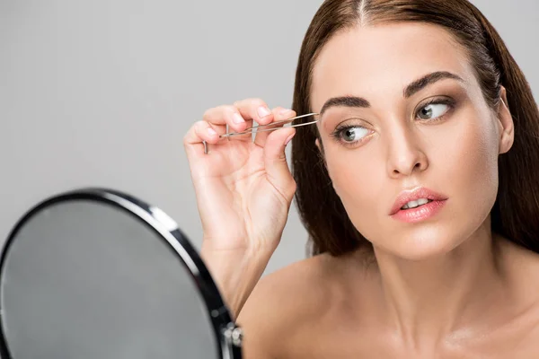 Young woman correcting shape of eyebrows and looking at mirror isolated on grey — Stock Photo