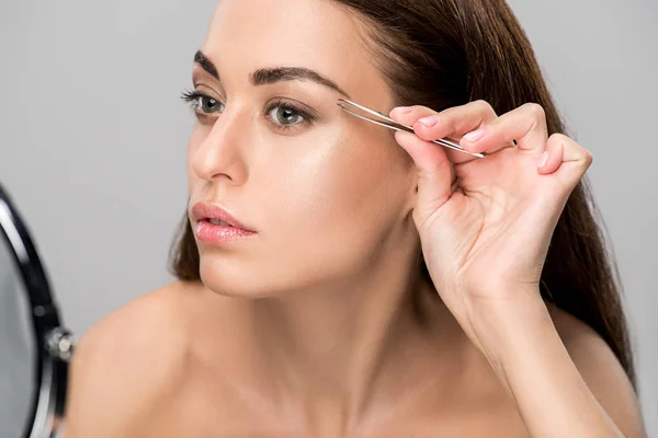 Belle jeune femme corrigeant la forme des sourcils avec des pinces isolées sur gris — Photo de stock