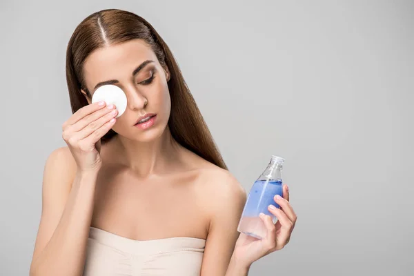 Attraktives Mädchen entfernt Make-up mit Baumwollscheibe und Pflegeprodukt, isoliert auf grau — Stockfoto