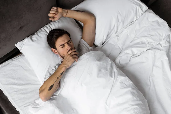 Draufsicht auf einen jungen Mann, der sich dehnt und gähnt, während er im Bett aufwacht — Stockfoto