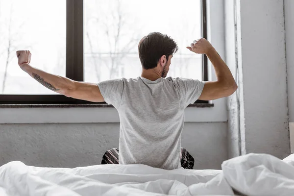 Rear view of young man in pajamas stretching while sitting in bed in the morning — Stock Photo