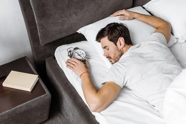 Vista de ángulo alto del joven sosteniendo el reloj despertador y durmiendo en la cama - foto de stock