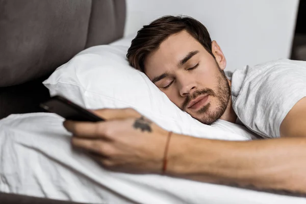 Bonito barbudo jovem dormindo na cama com smartphone na mão — Fotografia de Stock