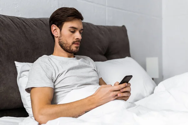 Guapo barbudo joven usando teléfono inteligente en la cama por la mañana — Stock Photo