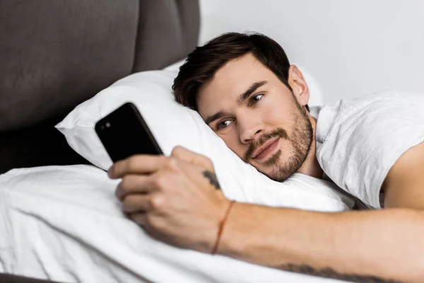 Bel homme barbu couché au lit et utilisant un smartphone — Photo de stock