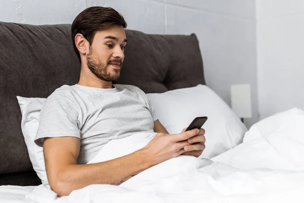 Handsome bearded man using smartphone in bed in the morning — Stock Photo