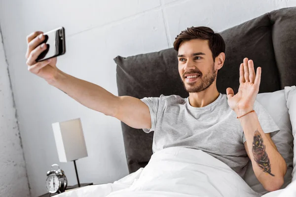 Smiling young man having video chat on smartphone and waving hand in bed — Stock Photo