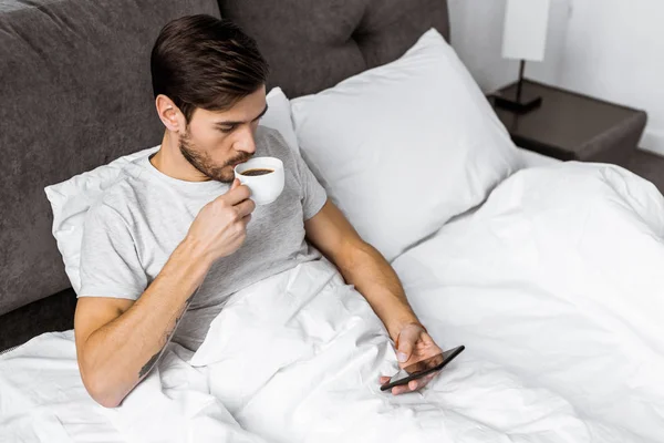 High angle view of young man using smartphone and drinking coffee in bed — Stock Photo