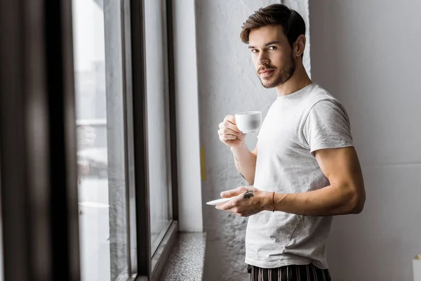 Schöner junger Mann mit einer Tasse Kaffee und Blick in die Kamera, während er morgens am Fenster steht — Stockfoto