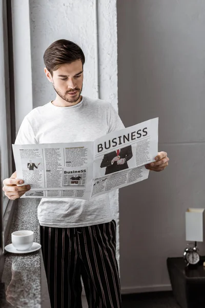 Красивый молодой человек в пижаме читает деловую газету дома — стоковое фото