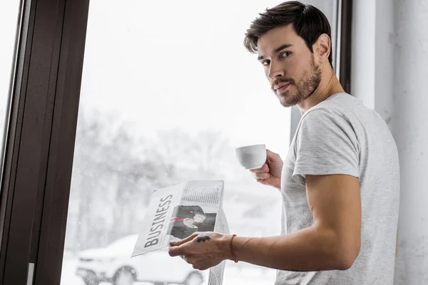 Joven guapo con taza de café y periódico mirando a la cámara mientras está de pie cerca de la ventana - foto de stock