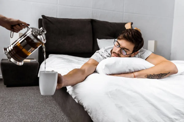 Plan recadré de quelqu'un versant du thé de théière tandis que l'homme endormi dans des lunettes couché sur le lit et tenant tasse — Photo de stock