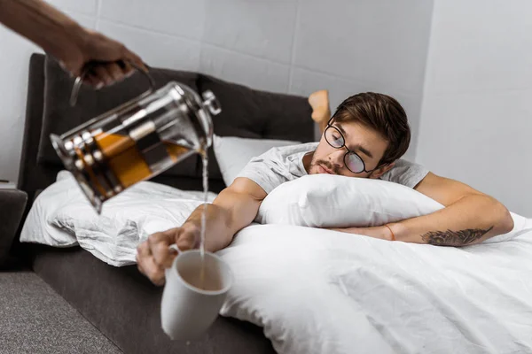 Partial view of someone pouring tea into cup while sleepy man in eyeglasses lying on bed — Stock Photo