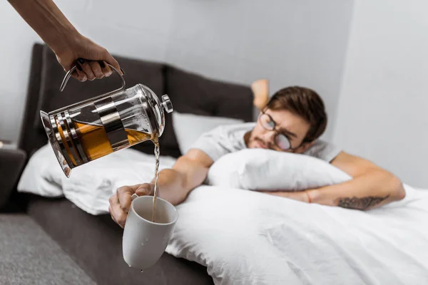 Vue partielle de quelqu'un versant du thé dans une tasse tout en dormant homme dans des lunettes couché sur le lit — Photo de stock