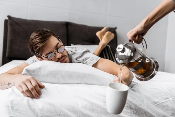 Cropped shot of someone pouring tea into cup while sleepy man in eyeglasses lying on bed — Stock Photo